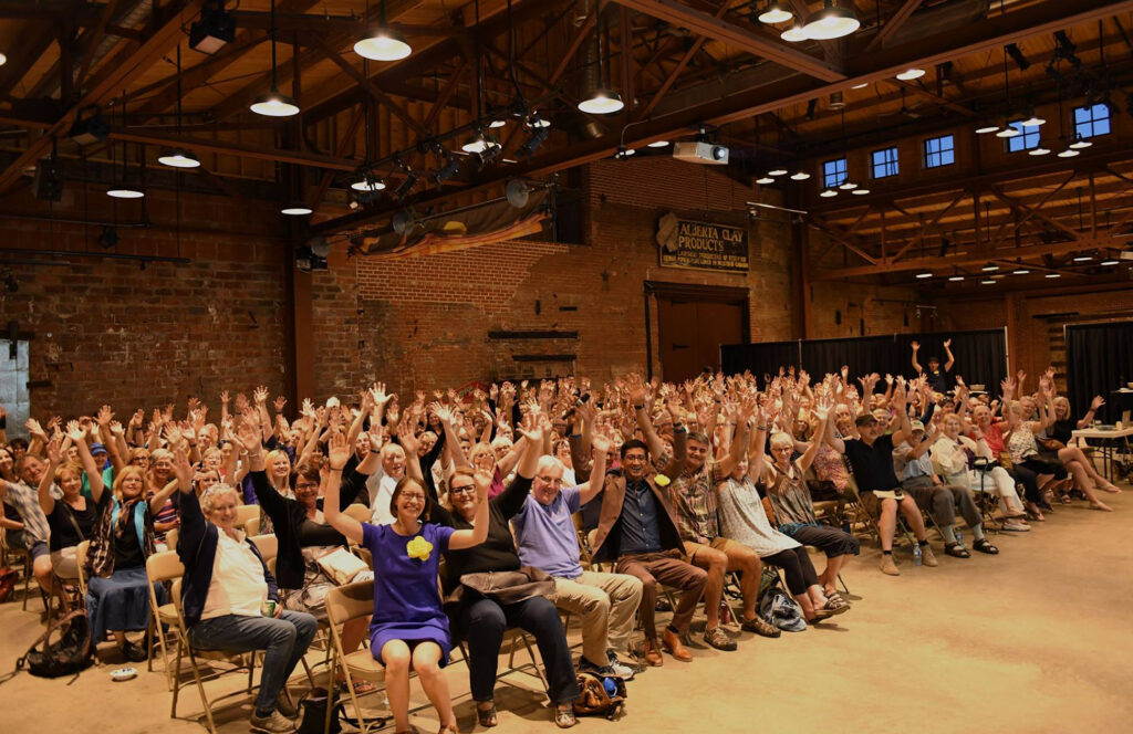 Large group of people sitting in a room with their hands raised in glee, during a fun presentation event