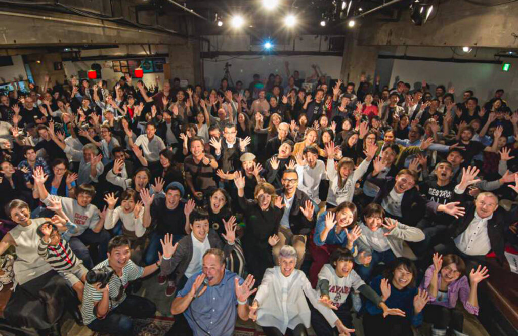 Large group of people sitting in a room with their hands raised in glee, during a fun presentation event